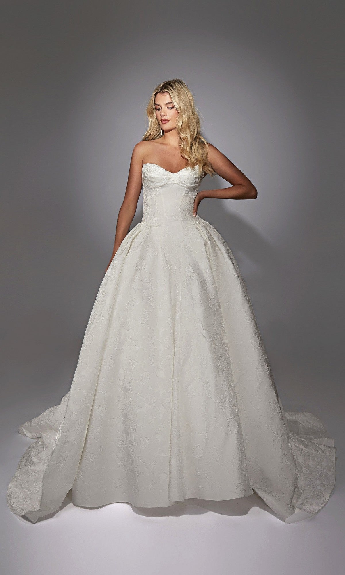 A woman wearing the Long White Dress: Alyce 7164, a strapless ball gown made of elegant floral jacquard, stands before a plain gray background.